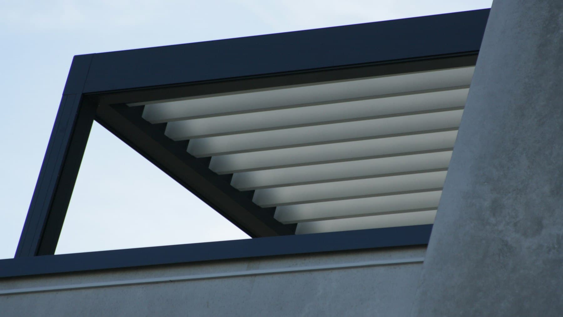 Dark aluminum louvered pergola roof against a blue sky beside a modern home