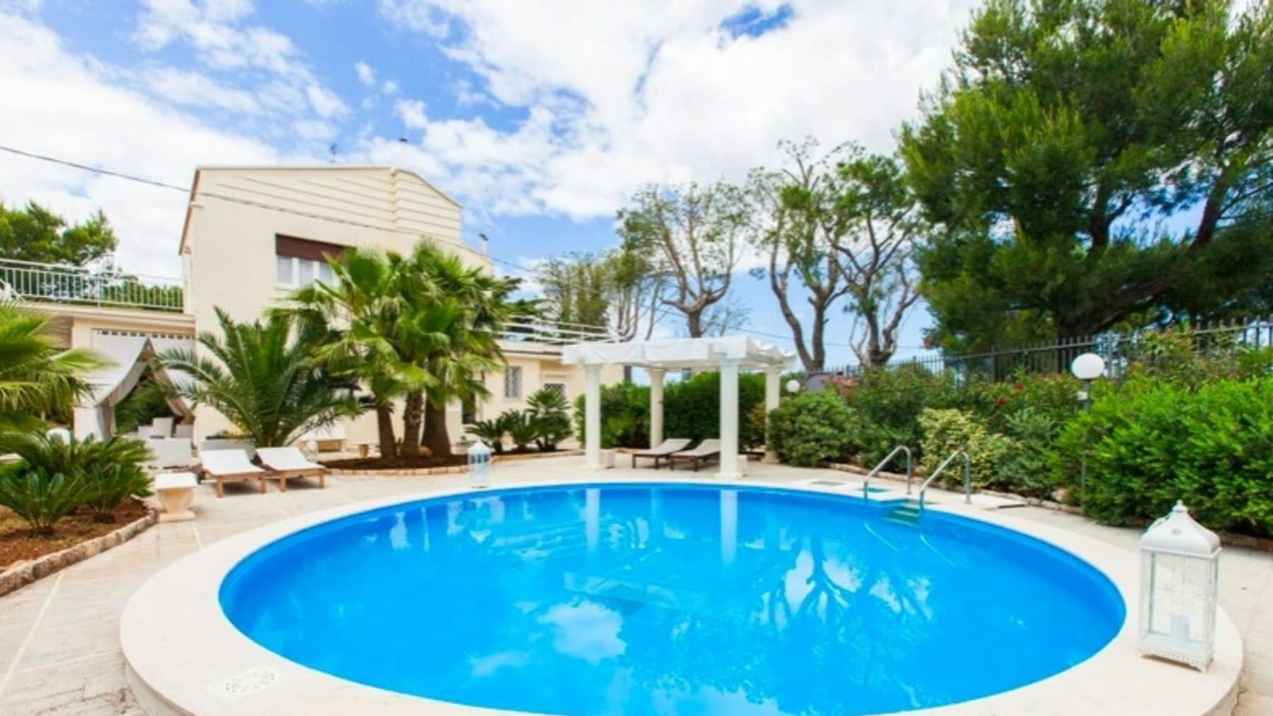 Round inground swimming pool surrounded by palm trees and a pergola in a landscaped backyard