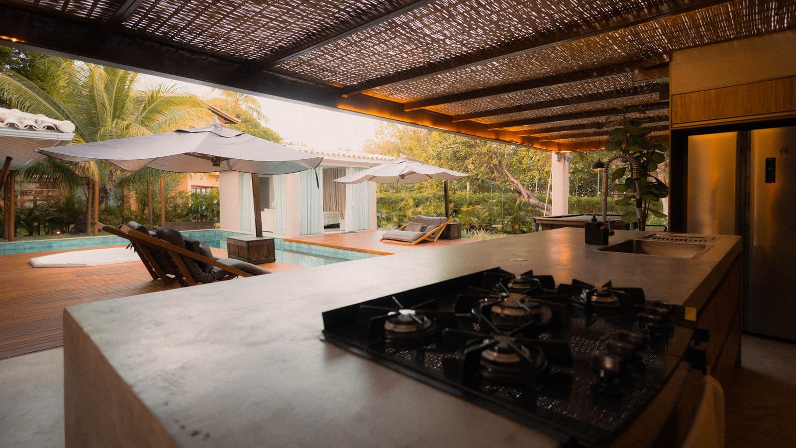 Resort-style outdoor kitchen island with a built-in gas cooktop overlooking a pool, loungers and tropical garden under a wood pergola