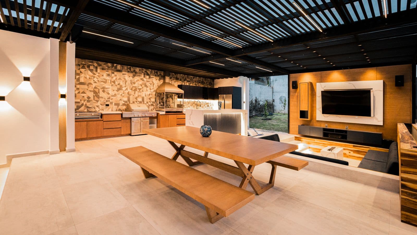Covered luxury outdoor kitchen and entertainment patio with built-in grill, wood dining table and mounted TV under a slatted pergola