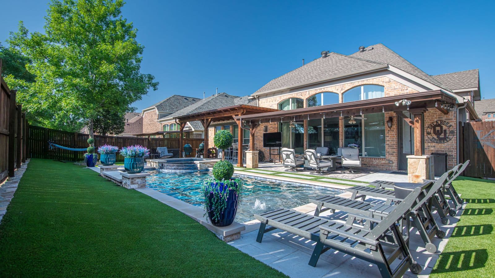 Upscale landscaped backyard with a modern swimming pool, deck loungers, and manicured artificial turf lawn beside a brick home