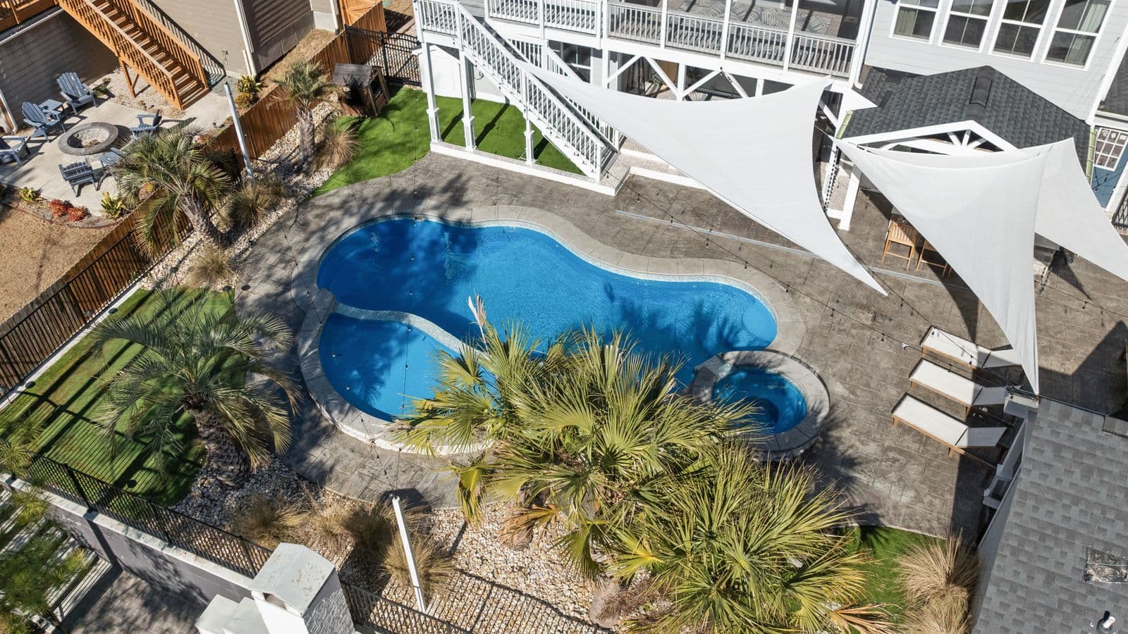 Aerial view of a luxury coastal backyard featuring a freeform pool bordered by a broad textured concrete pool-deck patio and palm trees