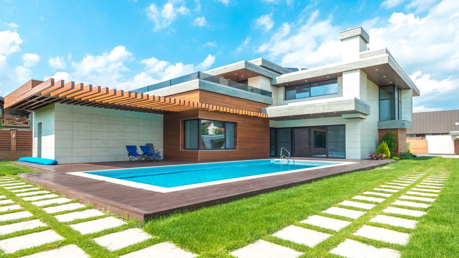 Modern concrete-and-wood two-story home with a rectangular backyard swimming pool, wood deck and paver-and-grass patio under a dramatic blue sky