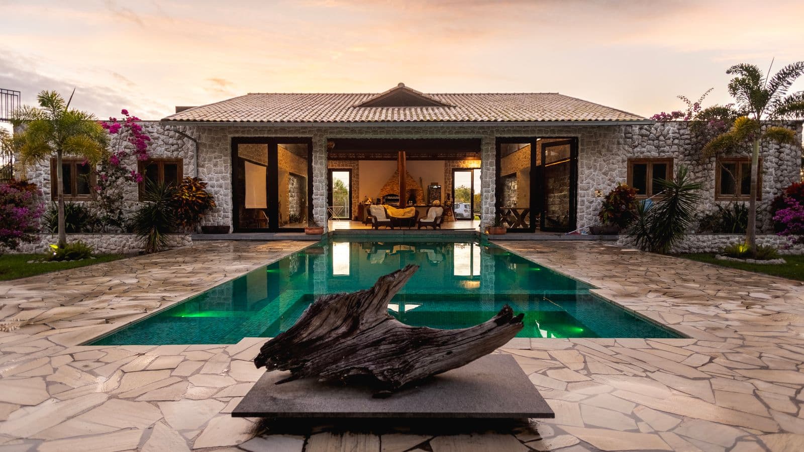 Stone luxury villa at sunset with a backyard swimming pool reflecting the house, flagstone patio, palms and bougainvillea