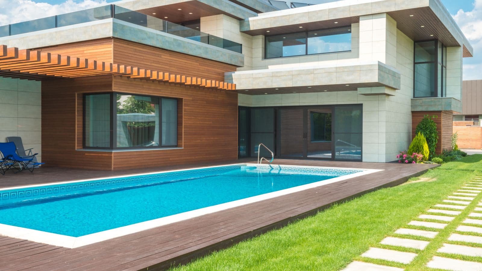 Contemporary luxury home with wood-clad facade beside a rectangular backyard swimming pool, wooden deck and manicured lawn with paver stepping stones