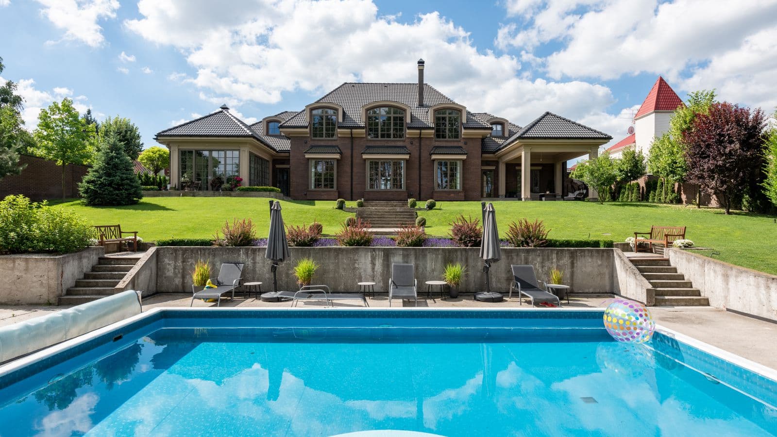 Luxury brick estate with a large in-ground blue swimming pool, poolside lounge chairs, umbrellas and manicured lawn under a bright sky
