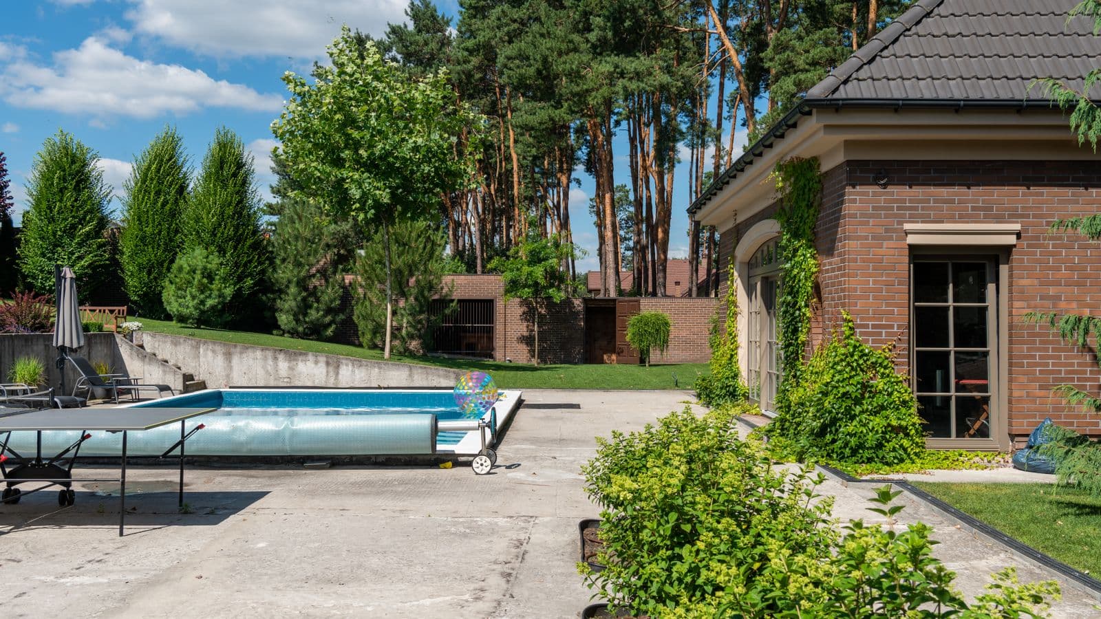 Sunny backyard with a poured-concrete pool-deck patio and driveway apron beside a brick home and manicured shrubs