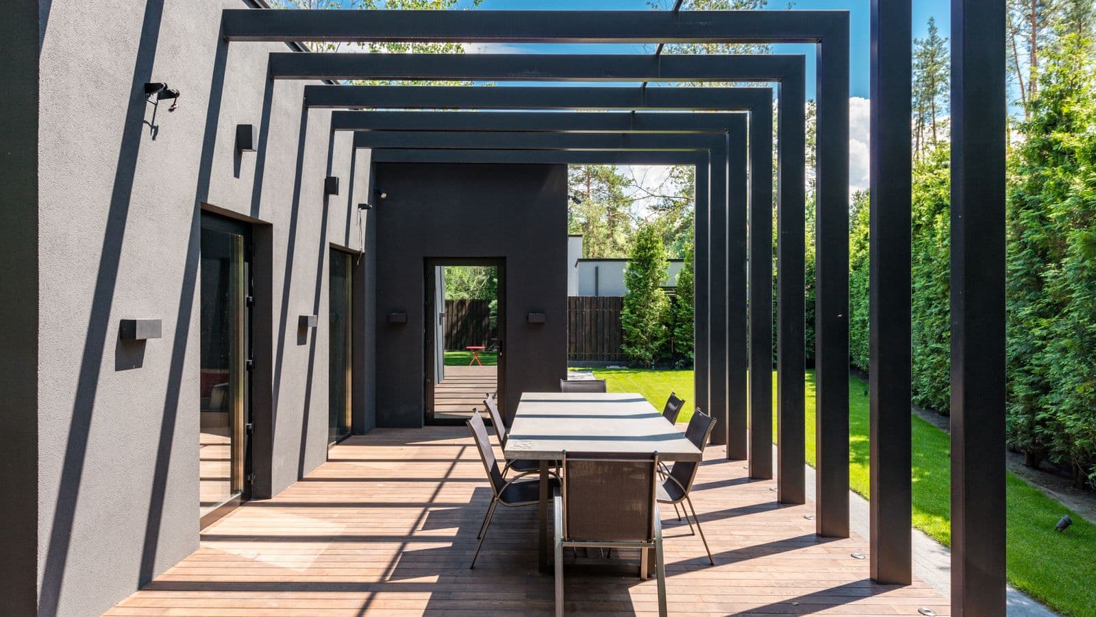 Modern black steel pergola casting geometric shadows over a wood-deck patio with an outdoor dining table beside a contemporary home
