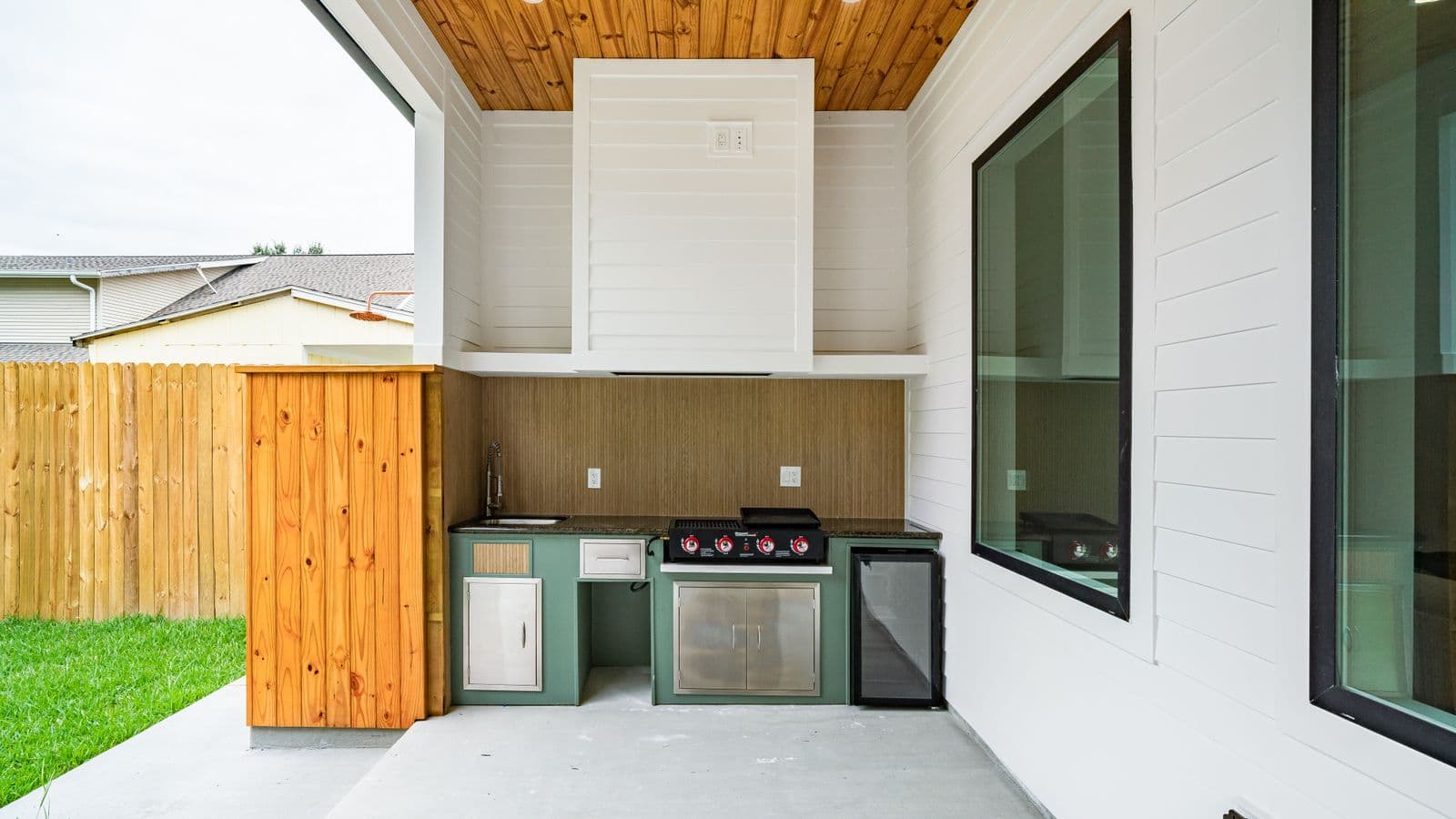 Modern built-in outdoor kitchen with stainless gas grill, mini-fridge, cabinetry and stone counter on a covered patio beside a lawn